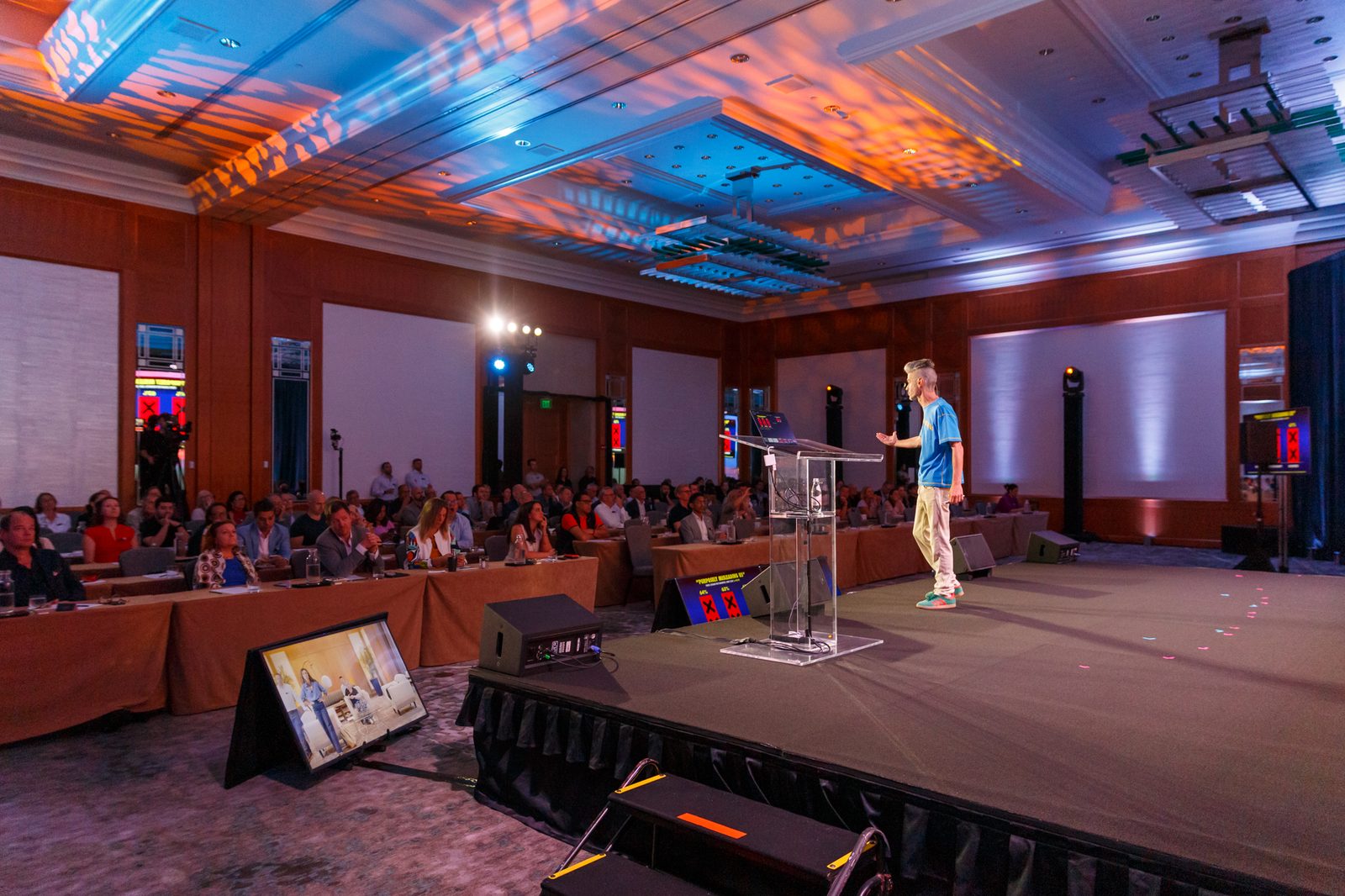 Speaker at a clear podium addressing a conference session in a hotel ballroom under blue and orange light