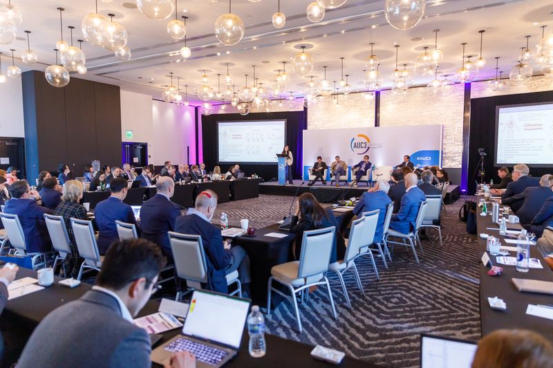 Hotel ballroom set for a working conference session, with attendees at long tables facing the stage
