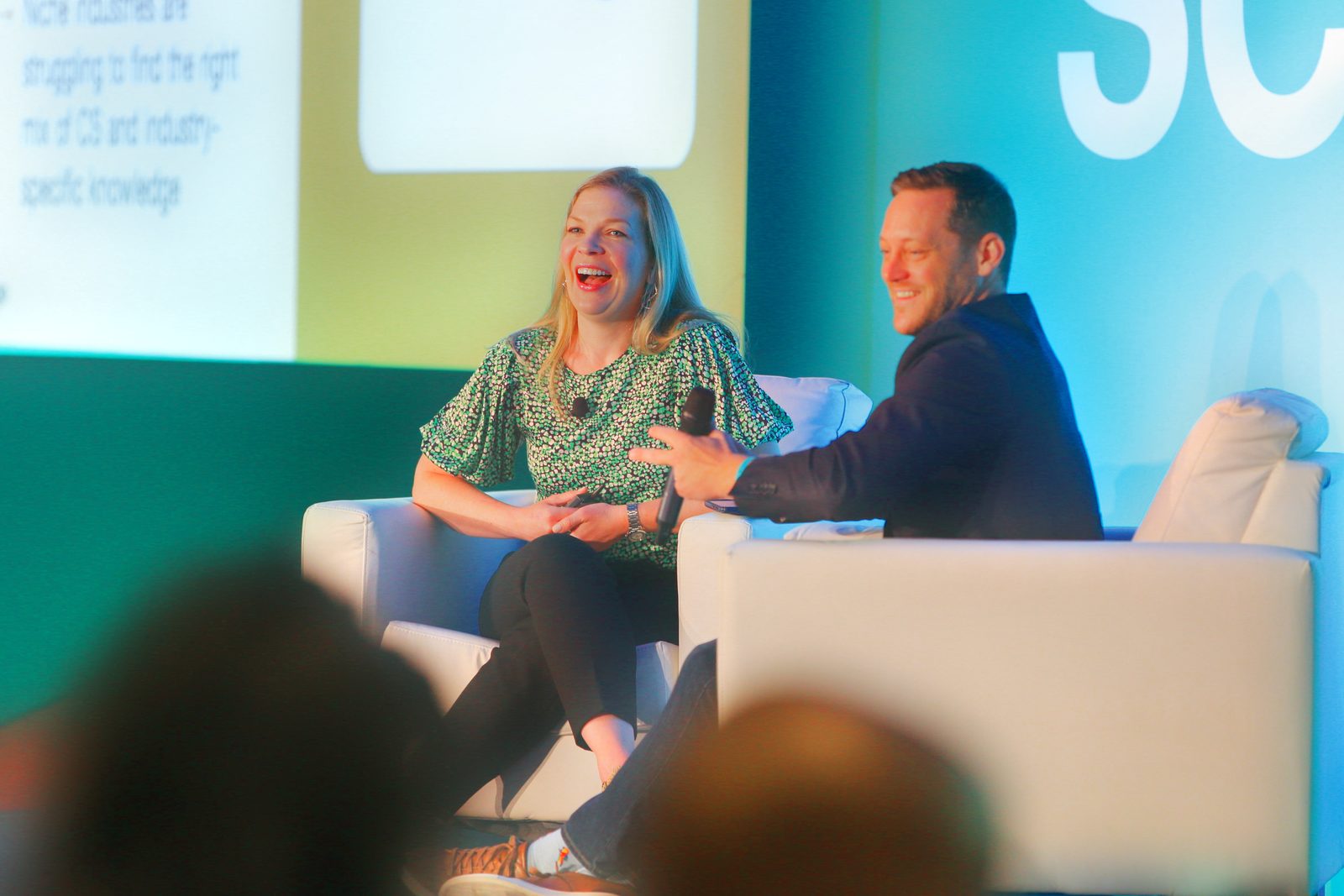 Two speakers laughing in armchairs on a conference stage, a slide and a brand wall behind them