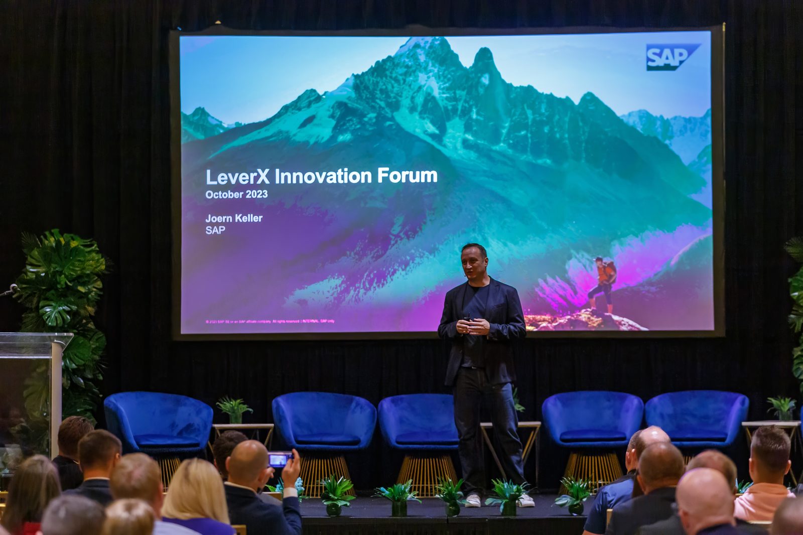 Speaker on stage at a technology forum in Miami, with blue lounge chairs and a full audience in front