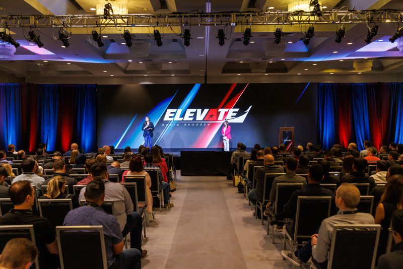 Full general session seen down the center aisle toward a stage with a giant event logo