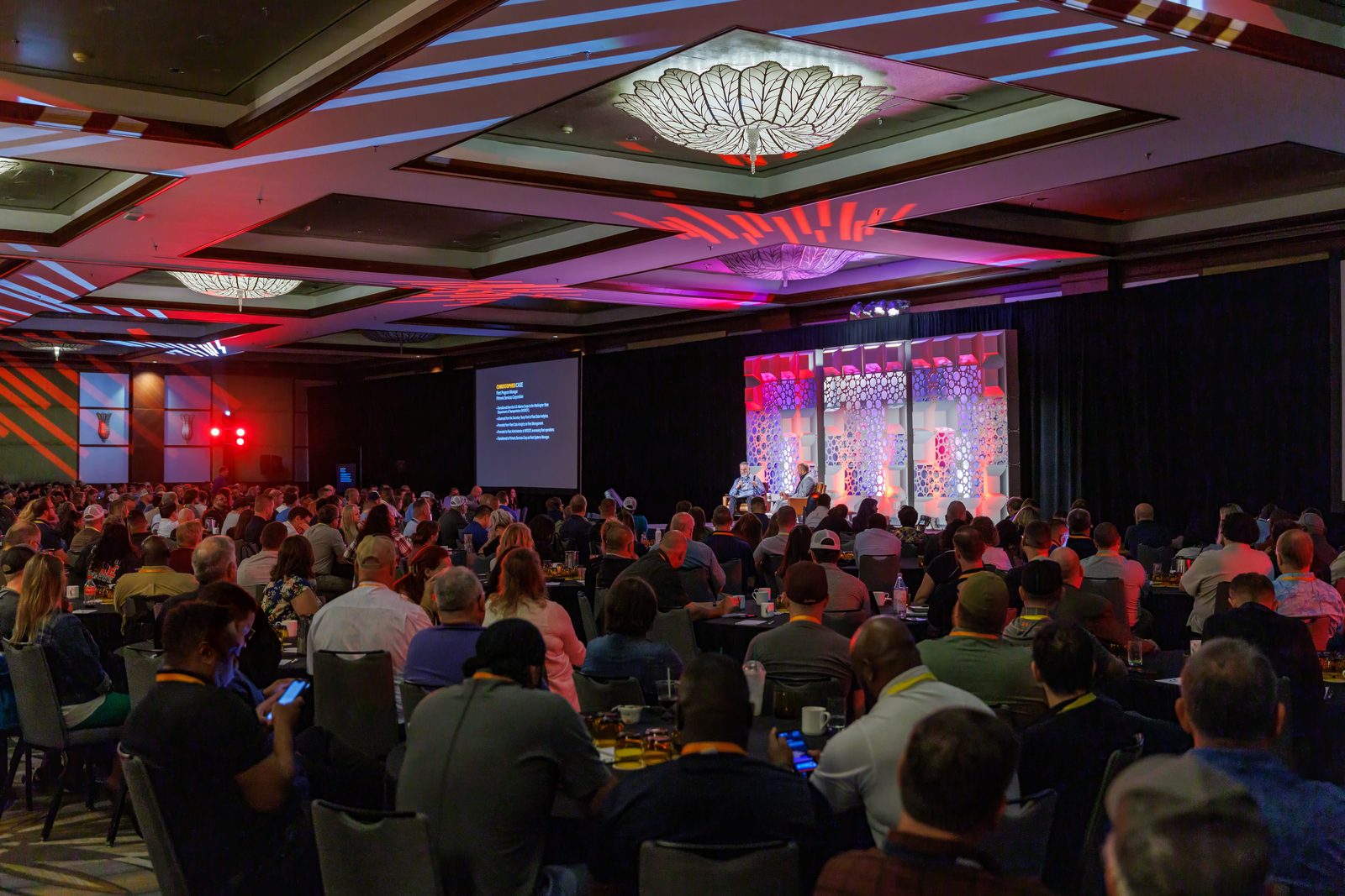 Packed hotel ballroom during a conference general session, with a lit stage set and screens on both sides