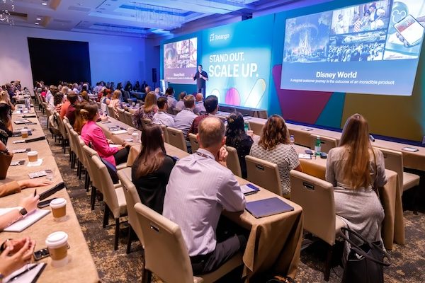 Packed conference general session with attendees at long tables facing large screens