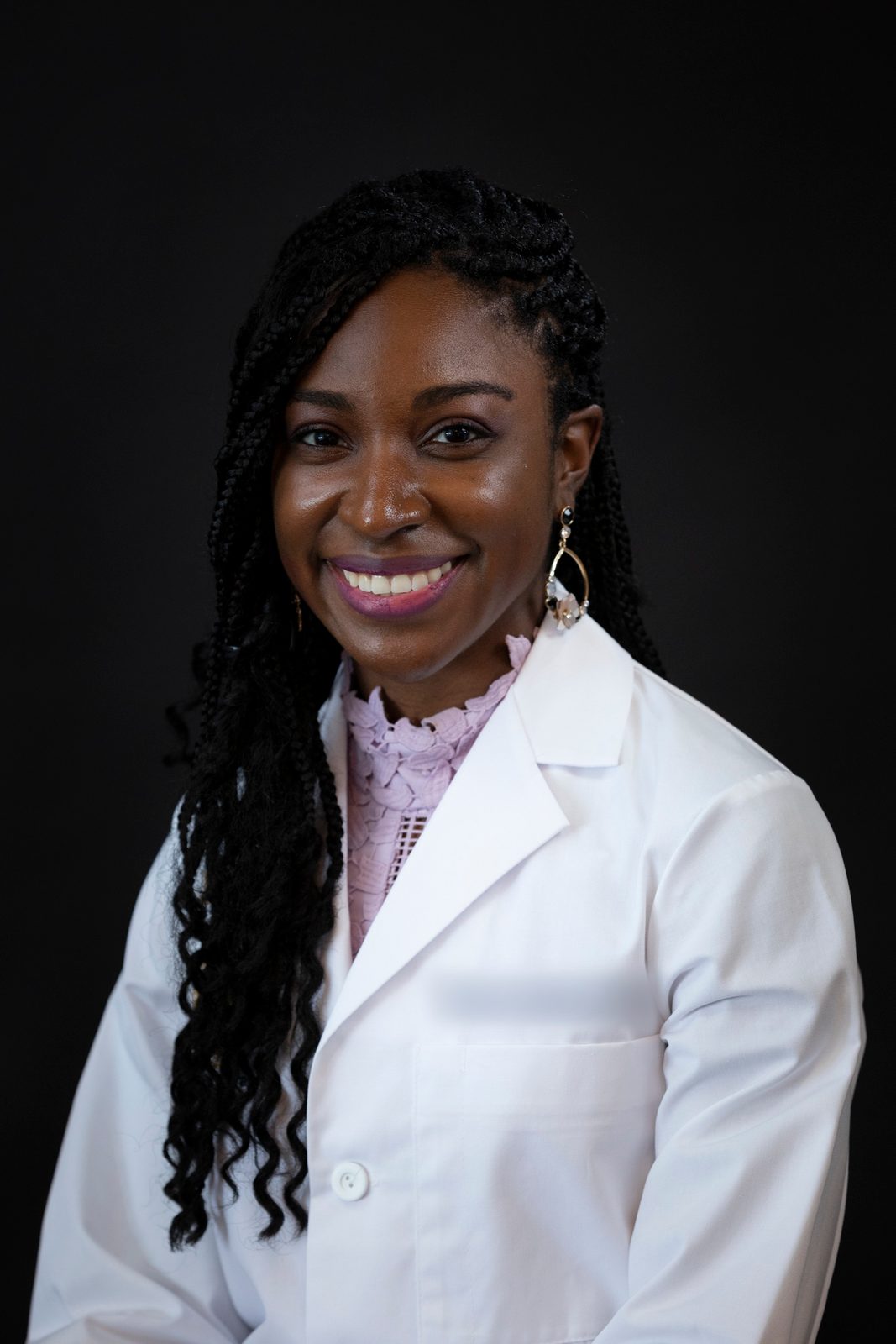 Headshot of a smiling woman with long braids in a white clinical coat against a black backdrop
