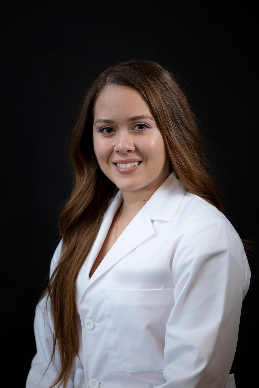 Headshot of a woman with long brown hair in a white clinical coat against a black backdrop