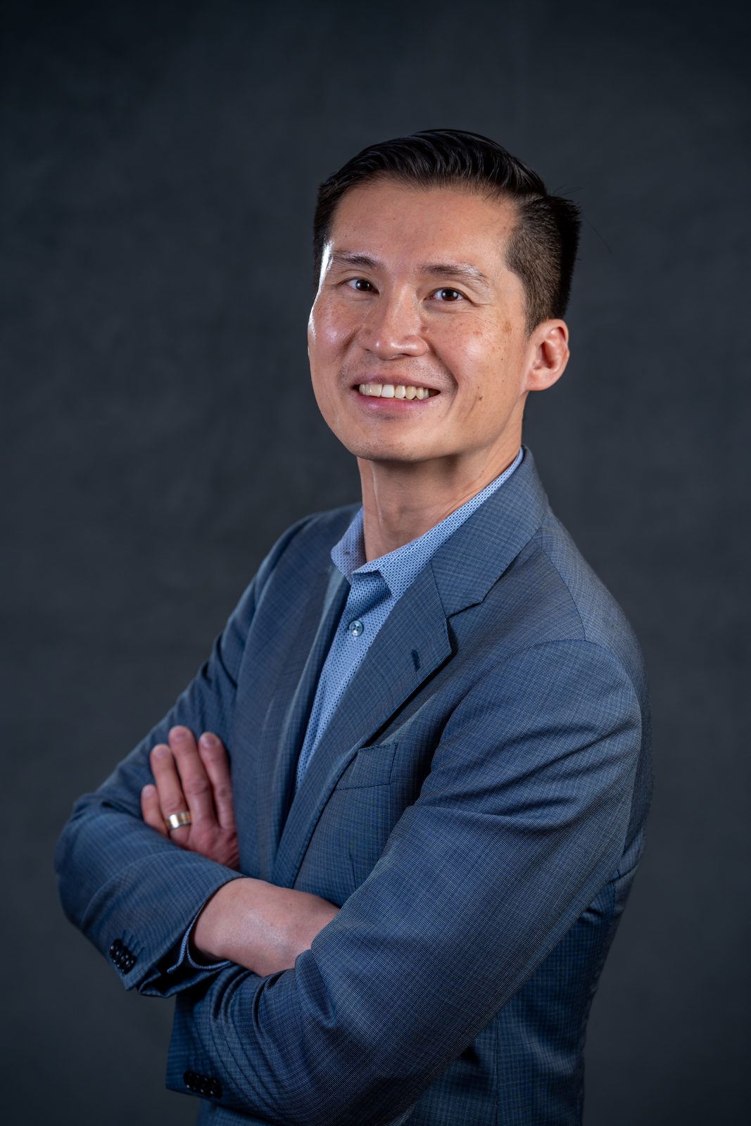 Headshot of a man in a blue suit with arms crossed against a gray backdrop