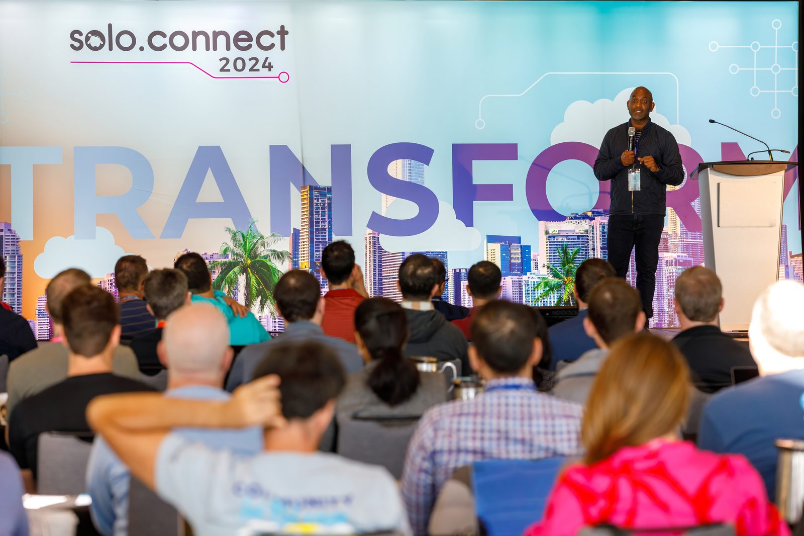 Keynote speaker on stage in front of a city-skyline backdrop, seen over a full room of attendees at a technology conference