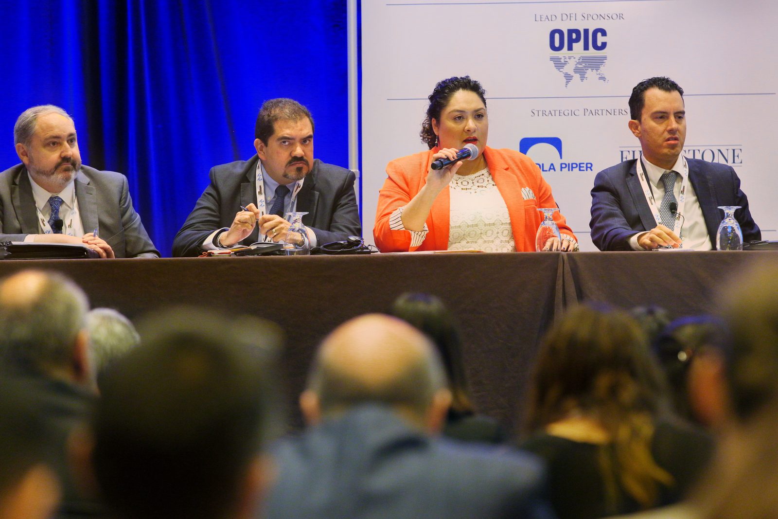 A panelist speaks into a microphone at a long table in front of a sponsor backdrop, the audience in the foreground
