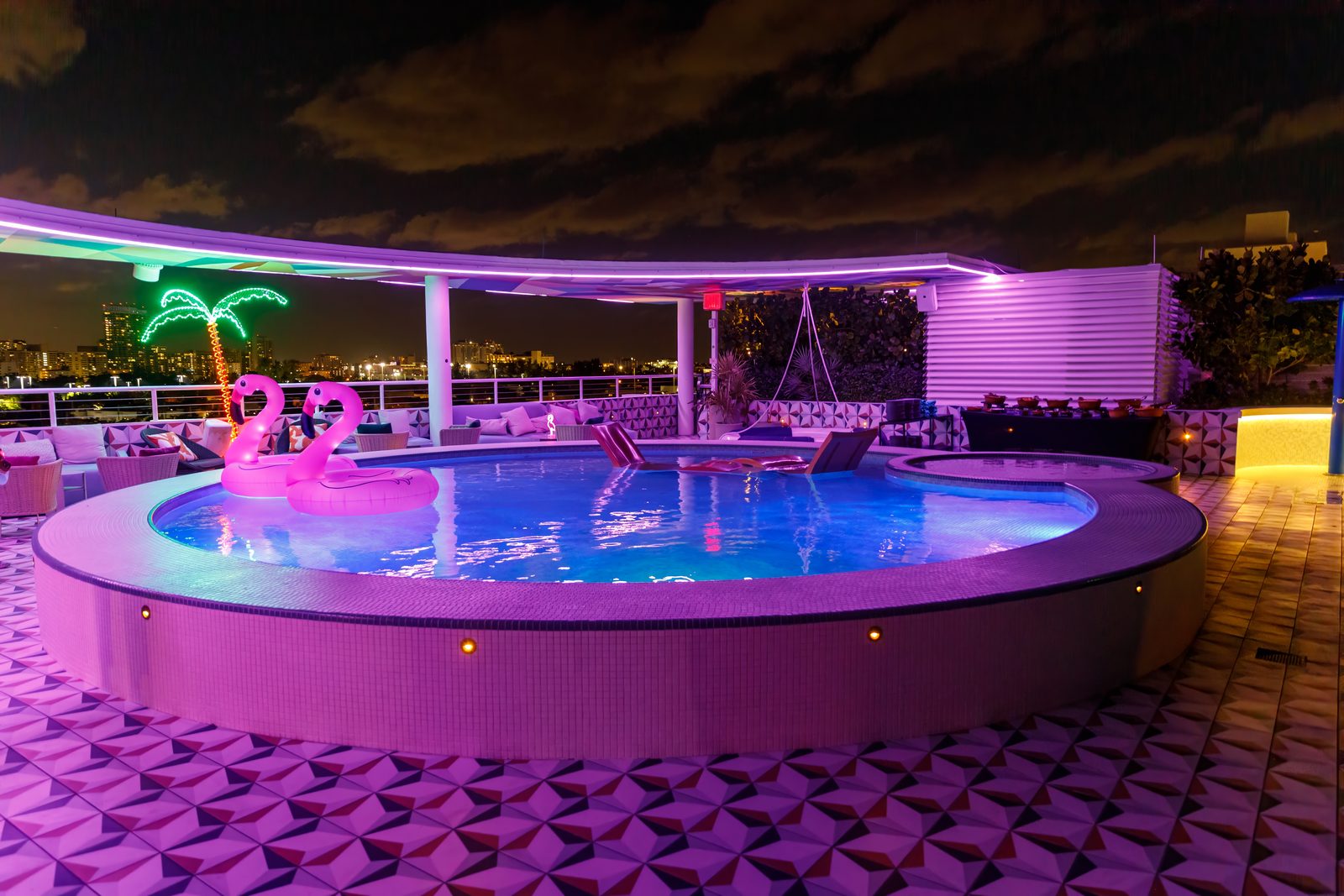 Rooftop pool lit purple at night with inflatable flamingos and neon palm trees