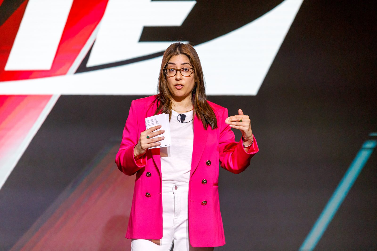 Speaker in a pink blazer presenting on a conference main stage in front of the event graphic