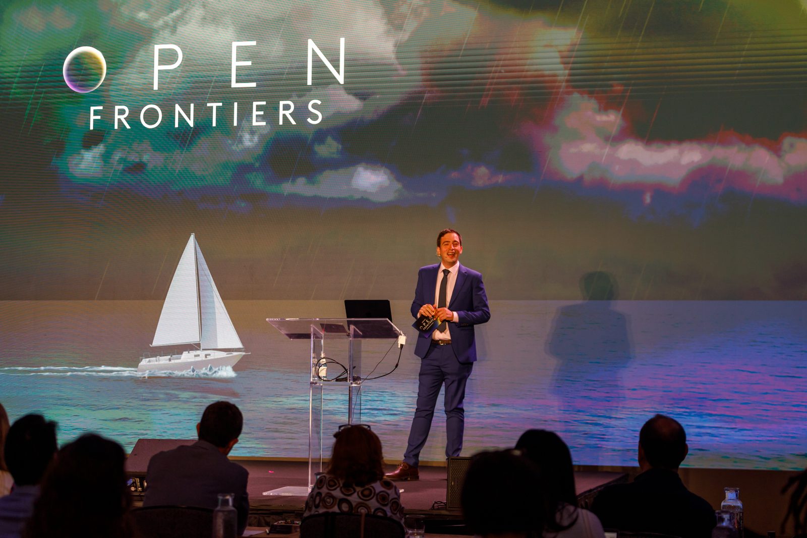Speaker on a conference stage in front of a large screen showing a sailboat on the sea at sunset, with the audience in silhouette