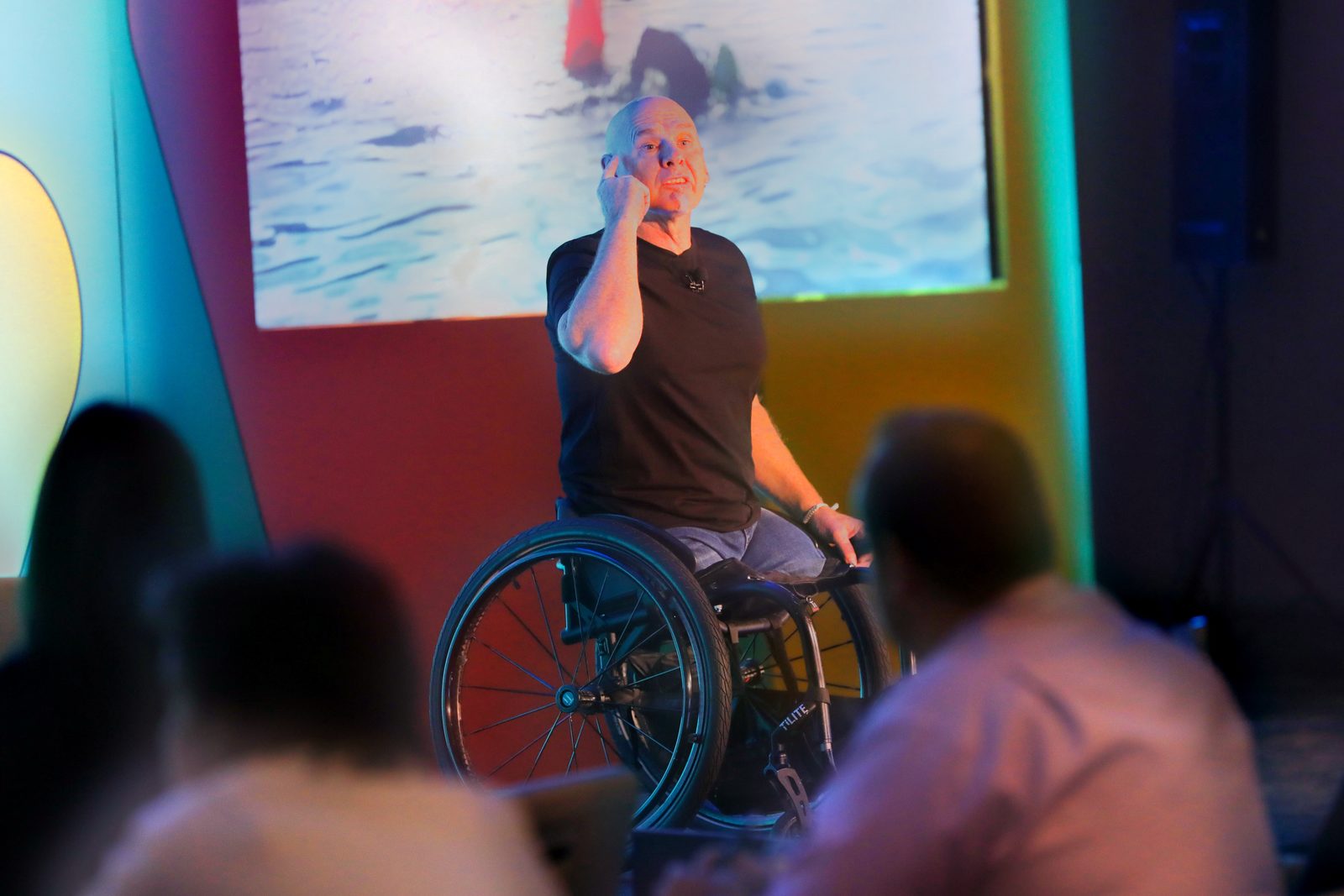 A speaker in a wheelchair talks to the audience in front of a projected ocean photo