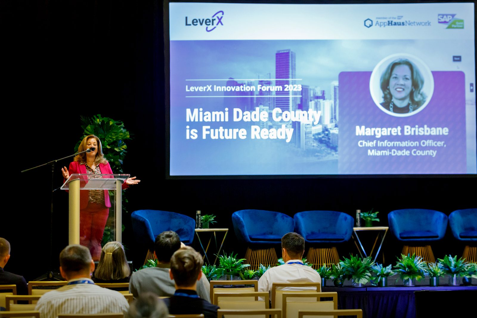 A speaker at the podium of a technology forum in Miami, with "Miami Dade County is Future Ready" on the screen behind her and attendees in the front rows